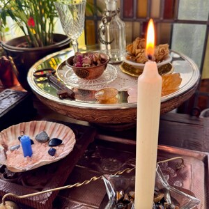 Op de afbeelding: Een brandende crèmekleurige dinerkaars staat in een stervormige glazen houder gevuld met tijgeroogstenen. Een schelp bevat roze zout en kleine donkere stenen, terwijl een zilveren dienblad een glazen fles, een sleutel en gedroogde bloemen toont.