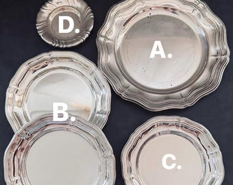 May include: A collection of four silver-toned metal plates and a small dish arranged on a dark surface. The items are labeled A, B, C, and D. Each piece features a smooth, reflective circular center and a decorative, scalloped, or wavy edge.