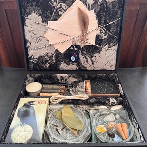 Op de afbeelding: Een zwarte doos met diverse spirituele items. Binnenin bevinden zich kaarsen, lucifers, een boek met een ravenillustratie en kleine glazen schalen met kruiden en kristallen. De doos is gevoerd met een zwart-wit patroonstof.