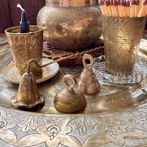 May include: A large round brass tray holding a brass bowl with etched floral patterns, two brass cups with leaf engravings, one containing a dark candle and the other matches, and three small brass bells of varying shapes and sizes.