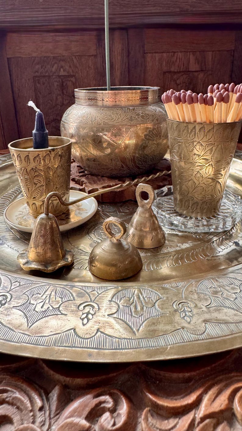 May include: A large round brass tray holding a brass bowl with etched floral patterns, two brass cups with leaf engravings, one containing a dark candle and the other matches, and three small brass bells of varying shapes and sizes.