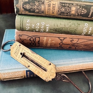 May include: A vintage brass pocket balance scale with a metal hook and a dual-scale face marked with Kilo and Zolle, resting atop a stack of four antique hardcover books in shades of teal, green, brown, and light blue with gold lettering.