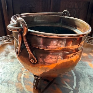 Puede incluir: Un caldero de cobre con cuerpo redondeado, tres patas pequeñas y dos asas grandes. La olla tiene una superficie brillante y reflectante y probablemente se utiliza para cocinar o con fines decorativos. El caldero está sobre una superficie de madera.