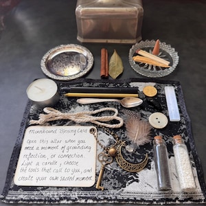 May include: A ritual kit on a black and white patterned cloth featuring a Moonbound Blessing Card, a brass crescent moon and key charm, a white tea light, black and gold candles, a silver spoon, a feather, two glass vials, and a selenite wand.
