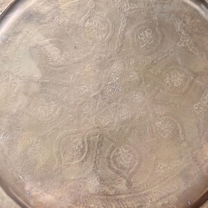 May include: A round brass tray featuring an intricate, hand-engraved floral and geometric pattern across its surface. The metal has a warm, aged patina with visible oxidation marks and a raised rim that frames the central decorative design.
