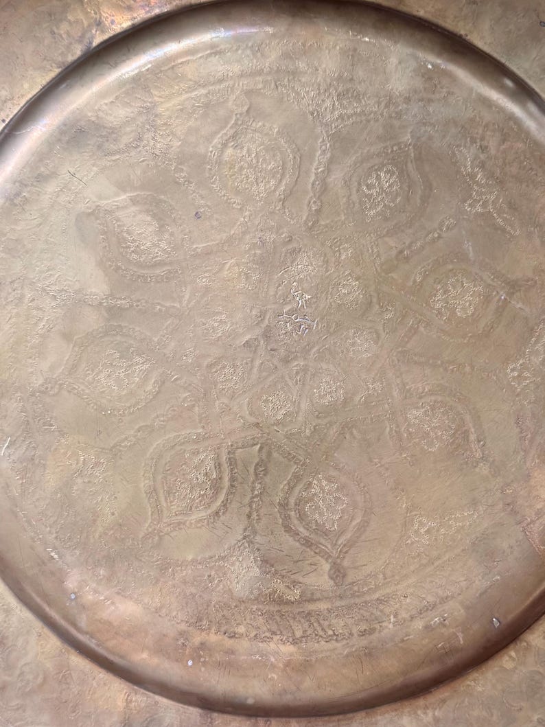 May include: A round brass tray featuring an intricate, hand-engraved floral and geometric pattern across its surface. The metal has a warm, aged patina with visible oxidation marks and a raised rim that frames the central decorative design.