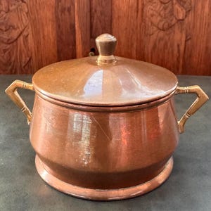 Puede incluir: Un recipiente redondo de cobre con tapa a juego y dos asas angulares de latón en los laterales. La tapa cuenta con un pequeño pomo central de latón. El cuerpo del recipiente tiene un acabado metálico liso con signos visibles de desgaste y pátina.