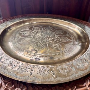 May include: A round brass tray featuring an intricate engraved geometric star pattern in the center and a decorative floral border along the rim. The metal surface displays a polished gold-toned finish with subtle signs of natural aging.