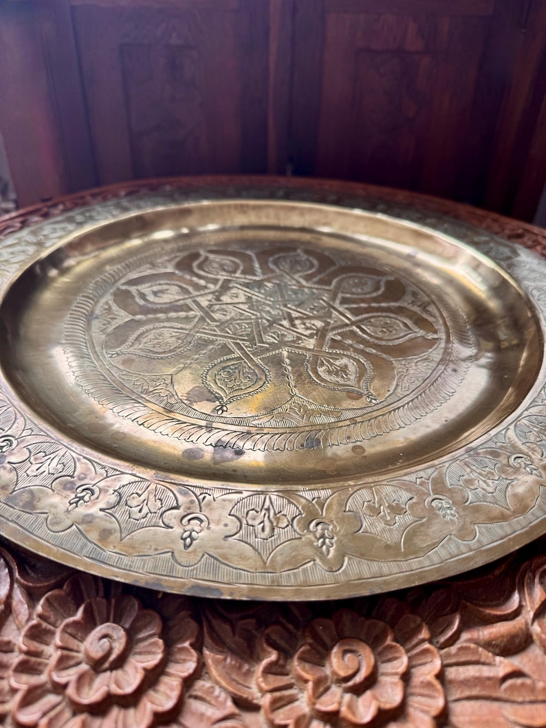 May include: A round brass tray featuring an intricate engraved geometric star pattern in the center and a decorative floral border along the rim. The metal surface displays a polished gold-toned finish with subtle signs of natural aging.