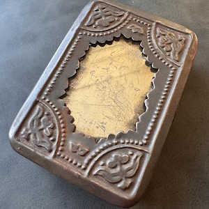 May include: An antique metal box, rectangular in shape, with a floral patterned border. The central recessed area has a gold-coloured surface. The metal has a tarnished, aged appearance, indicating it is vintage.
