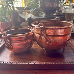 May include: Two antique copper pots with handles and a small copper bucket sit on a wooden tray. The pots have a rich, reddish-brown color and are likely used for decorative purposes. The background includes plants and a window.