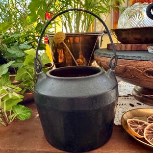 Op de afbeelding: Een matzwarte gietijzeren pot met een brede, ronde vorm en een stevig, gebogen metalen handvat dat aan twee lussen aan de zijkant is bevestigd. De pot heeft een dik, getextureerd oppervlak en een brede, open cirkelvormige bovenkant.