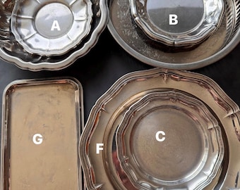 May include: A collection of antique silver serving pieces, including decorative trays and plates. The items have ornate edges and varying sizes. The arrangement includes a rectangular tray and several round plates and bowls, all with a reflective surface.