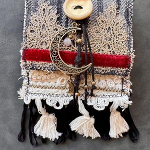 Pode incluir: Uma bolsa têxtil artesanal com um fundo semelhante ao denim, adornada com renda dourada, uma faixa de veludo vermelho e franjas brancas. Apresenta um pingente de lua crescente, um botão grande e borlas penduradas, criando uma estética boêmia.