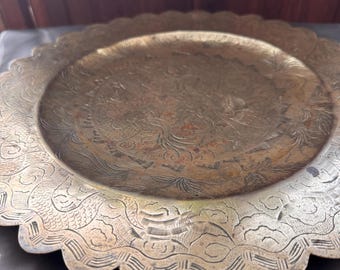 May include: A detailed, antique brass plate with an ornate, embossed design. The plate has a scalloped edge and a circular, raised center. The intricate patterns include floral and wave-like motifs, creating a vintage aesthetic. The plate is likely made of brass.