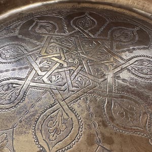 May include: A large, round, metallic brass tray featuring an intricate, hand-engraved geometric star pattern at its center, surrounded by detailed floral motifs and a decorative border along the raised outer rim with a weathered patina finish.