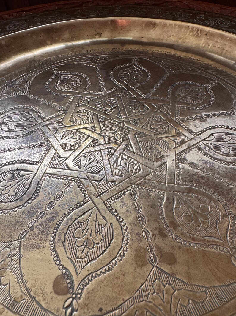 May include: A large, round, metallic brass tray featuring an intricate, hand-engraved geometric star pattern at its center, surrounded by detailed floral motifs and a decorative border along the raised outer rim with a weathered patina finish.