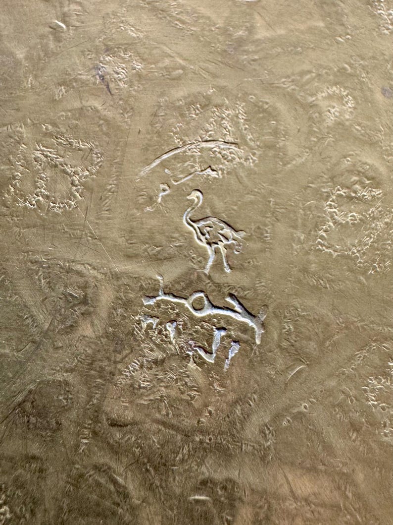 May include: A textured brass surface featuring an engraved design of a bird standing between two lines of Arabic script. The metal has a dull, aged gold finish with visible scratches and uneven markings across the entire metallic plate.