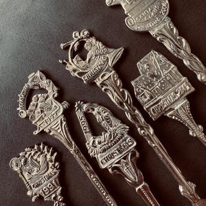 Op de afbeelding: Een verzameling van vijf zilverkleurige souvenirlepels met gedetailleerde kerstontwerpen. Elk lepeltje heeft een unieke reliëfscène op de steel, waaronder een kerststal, een huis en de kerstman, met jaartallen zoals 1991, 1992, 1995, 2002, 2003 en 2004 erin gegraveerd.