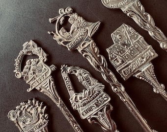 May include: A collection of five silver-toned metal souvenir spoons featuring intricate holiday-themed designs. Each spoon handle displays a unique embossed scene, including a nativity, a house, and Santa, with years such as 1991, 1992, 1995, 2002, 2003, and 2004 engraved.