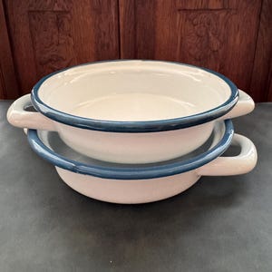 Op de afbeelding: Twee gestapelde ronde geëmailleerde ovenschaaltjes met een crèmekleurige binnen- en buitenkant, voorzien van een donkerblauwe rand en twee handgrepen aan de zijkanten van elk schaaltje. De schaaltjes hebben een gladde, glanzende afwerking.