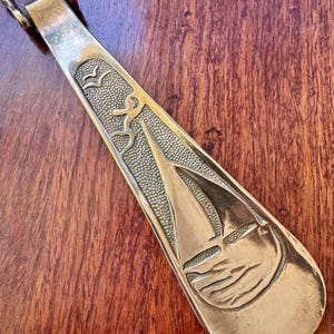 May include: A brass shoe horn with a sailboat design. The shoe horn has a curved handle and a long, flat body. The sailboat is depicted with a detailed sail and waves. The design also includes birds and a textured background.