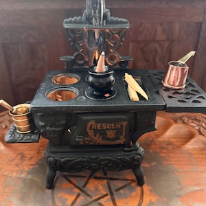 Pode incluir: Um fogão em miniatura de ferro fundido preto com a palavra CRESCENT em relevo na porta frontal. O fogão apresenta um pequeno caldeirão preto com um cone de incenso fumegante, duas aberturas circulares, um pedaço de madeira e duas pequenas panelas de metal.