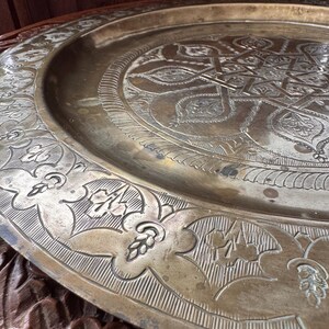 May include: A large, circular brass tray featuring intricate, hand-engraved geometric and floral patterns. The center displays a complex star motif, while the wide, raised rim is decorated with repeating leaf and petal designs in a metallic gold tone.