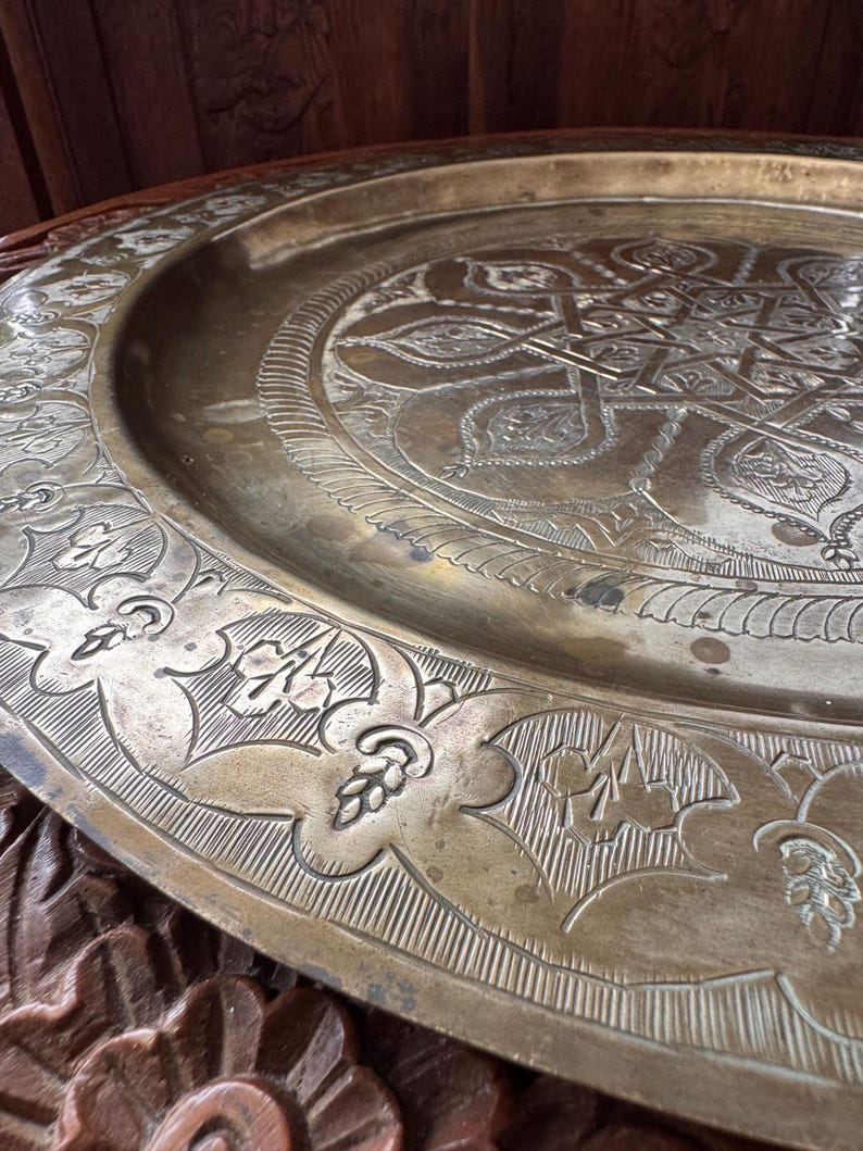 May include: A large, circular brass tray featuring intricate, hand-engraved geometric and floral patterns. The center displays a complex star motif, while the wide, raised rim is decorated with repeating leaf and petal designs in a metallic gold tone.