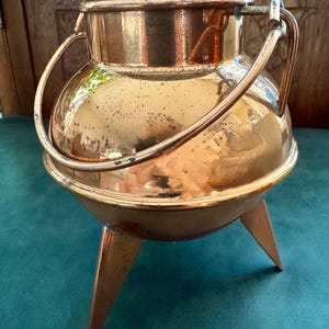 Pode incluir: Uma panela de cobre polido com corpo arredondado, uma ampla abertura circular e uma alça resistente fixada nas laterais. O recipiente é sustentado por três pernas pontiagudas que se estendem da base, com uma superfície metálica reflexiva.