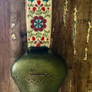 Può includere: Un campanaccio in metallo verde con la scritta BURGGRABENKLAMM stampata sul davanti. La campana è fissata a un cinturino in tessuto intrecciato con un motivo floreale ripetuto nei colori rosso, verde acqua e viola su sfondo crema.