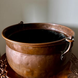Puede incluir: Una olla redonda de cobre martillado con una parte superior ancha y abierta y un borde liso reforzado. La olla presenta una pátina oscura y desgastada, y está equipada con dos asas laterales de metal para un mango, con forma profunda.