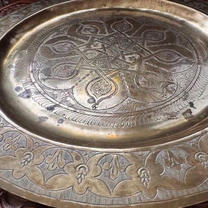 May include: A large, round brass tray featuring intricate hand-engraved geometric and floral patterns. The center displays a complex star-shaped motif surrounded by detailed filigree, while the wide rim is decorated with repeating leaf designs.