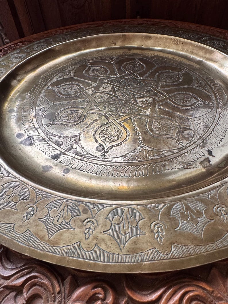 May include: A large, round brass tray featuring intricate hand-engraved geometric and floral patterns. The center displays a complex star-shaped motif surrounded by detailed filigree, while the wide rim is decorated with repeating leaf designs.