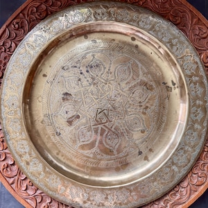 May include: A round brass tray featuring an intricate engraved geometric star pattern in the center and a decorative floral border. The tray is positioned on top of a circular wooden base with a carved floral relief design along the edge.