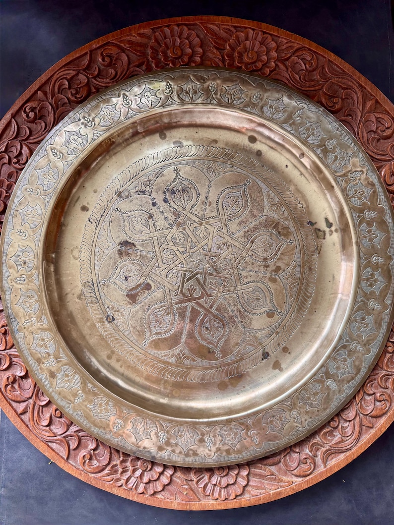 May include: A round brass tray featuring an intricate engraved geometric star pattern in the center and a decorative floral border. The tray is positioned on top of a circular wooden base with a carved floral relief design along the edge.