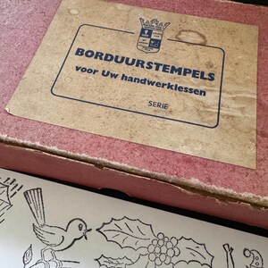 Könnte beinhalten: Ein rechteckiger rosa Pappkarton mit einem beigen Etikett, das den Text BORDUURSTEMPELS voor Uw handwerklessen SERIE und ein Wappen zeigt. Unter dem Karton liegt ein weißes Blatt mit schwarzen Stempelmotiven von einem Vogel, Stechpalme und einer Figur.