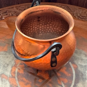 Puede incluir: Un pequeño caldero de cobre martillado con un asa de metal oscuro. El caldero tiene un cuerpo redondeado y un interior texturizado. El asa está sujeta con correas de cuero. El cobre tiene un tono naranja cálido.