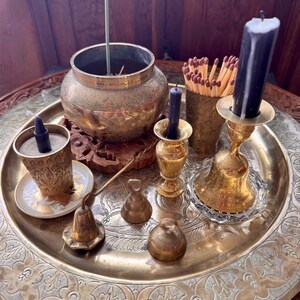 May include: A large round brass tray holding a collection of brass items including an etched bowl with an incense stick, a small cup with a dark candle, a match holder with wooden matches, a tall candle holder with a dark candle, and three small bells.