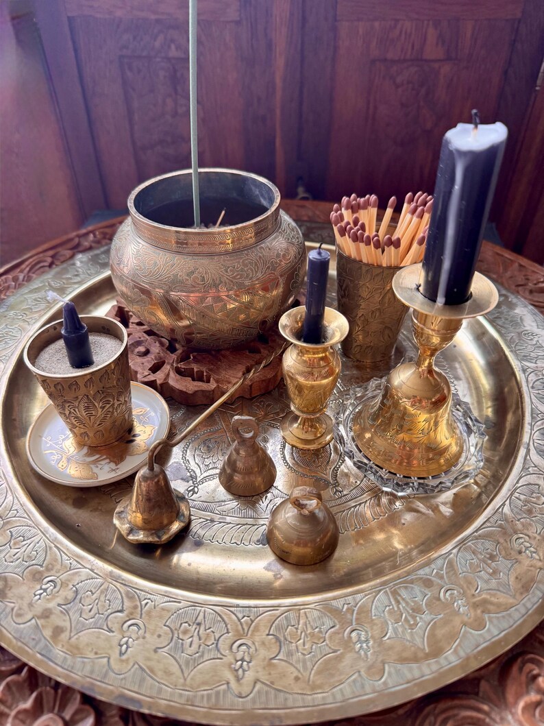 May include: A large round brass tray holding a collection of brass items including an etched bowl with an incense stick, a small cup with a dark candle, a match holder with wooden matches, a tall candle holder with a dark candle, and three small bells.