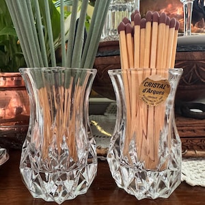 Op de afbeelding: Twee heldere, facetgeslepen glazen vazen. Eén vaas bevat groene wierookstokjes, de andere houten lucifers met rode uiteinden. De lucifers zitten in een vaas met een "Cristal d'Arques" sticker.