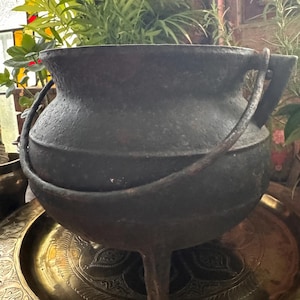 Puede incluir: Un caldero de hierro fundido oscuro y pesado con cuerpo redondeado, tres patas cortas y un asa de metal curvada unida a los lados. La superficie de la olla tiene un acabado mate y texturizado, y el borde es ancho con una forma ligeramente acampanada.