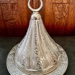 Puede incluir: Un plato de servicio de metal color plata con una tapa cónica que presenta un intrincado patrón floral en relieve y un remate en forma de media luna en la parte superior. La base es un plato circular con un borde floral grabado.
