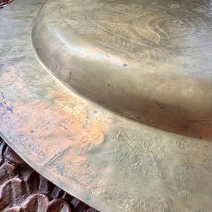 May include: A large, circular brass tray featuring an engraved, intricate pattern across its surface. The metal has a weathered, golden-brown patina with visible surface scratches and signs of age, showing a raised central section.