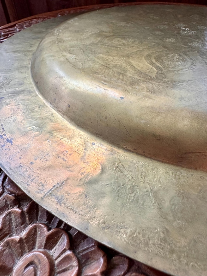 May include: A large, circular brass tray featuring an engraved, intricate pattern across its surface. The metal has a weathered, golden-brown patina with visible surface scratches and signs of age, showing a raised central section.