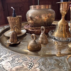May include: A collection of brass items including a large engraved tray, a round bowl with intricate floral patterns, a tall candlestick with a black candle, a small cup with a black candle, and three small brass bells of varying shapes and sizes.