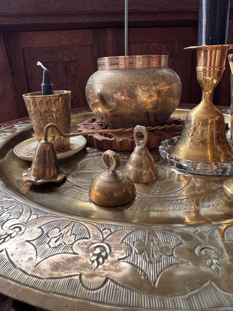 May include: A collection of brass items including a large engraved tray, a round bowl with intricate floral patterns, a tall candlestick with a black candle, a small cup with a black candle, and three small brass bells of varying shapes and sizes.