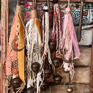 Può includere: Cinque ornamenti decorativi appesi composti da nastri stratificati, pizzo e spago. Ogni ornamento include campanelli in metallo color bronzo a forma di ghianda, piccole perline e ciondoli a forma di mezzaluna e stella, sospesi da cappi di spago.