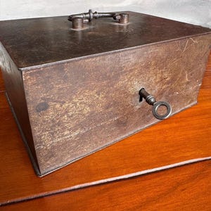 May include: A vintage metal box with a key in the lock and a handle on top. The box has a dark, aged appearance with visible wear and tear. The key is inserted into the keyhole on the side. The box is sitting on a wooden surface.