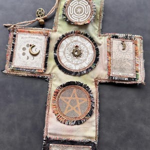 Puede incluir: Colgante de pared hecho a mano en forma de cruz con varios símbolos celestiales y ocultos. Presenta un diseño central de explosión solar, un pentagrama e imágenes de fases lunares. La cruz está hecha de tela con bordes deshilachados y una cuerda de yute para colgar.
